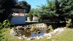 Ein kleiner Teich, umgeben von Felsen, ein weißes Gebäude mit Ziegeldach und Bäume im Hintergrund.