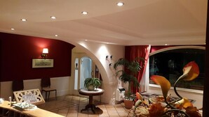 Ein Hotelzimmer mit roter Wand, einem runden Tisch mit Lampe, einem Stuhl, einer Topfpflanze und einem Fenster mit Blick auf den Nachthimmel.