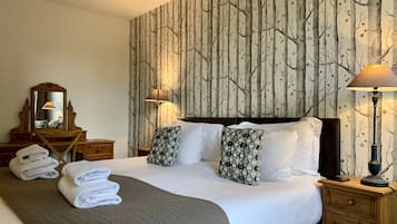 A neatly made bed with white linens and patterned pillows, a wooden nightstand with a lamp, and a wall with a tree pattern wallpaper.