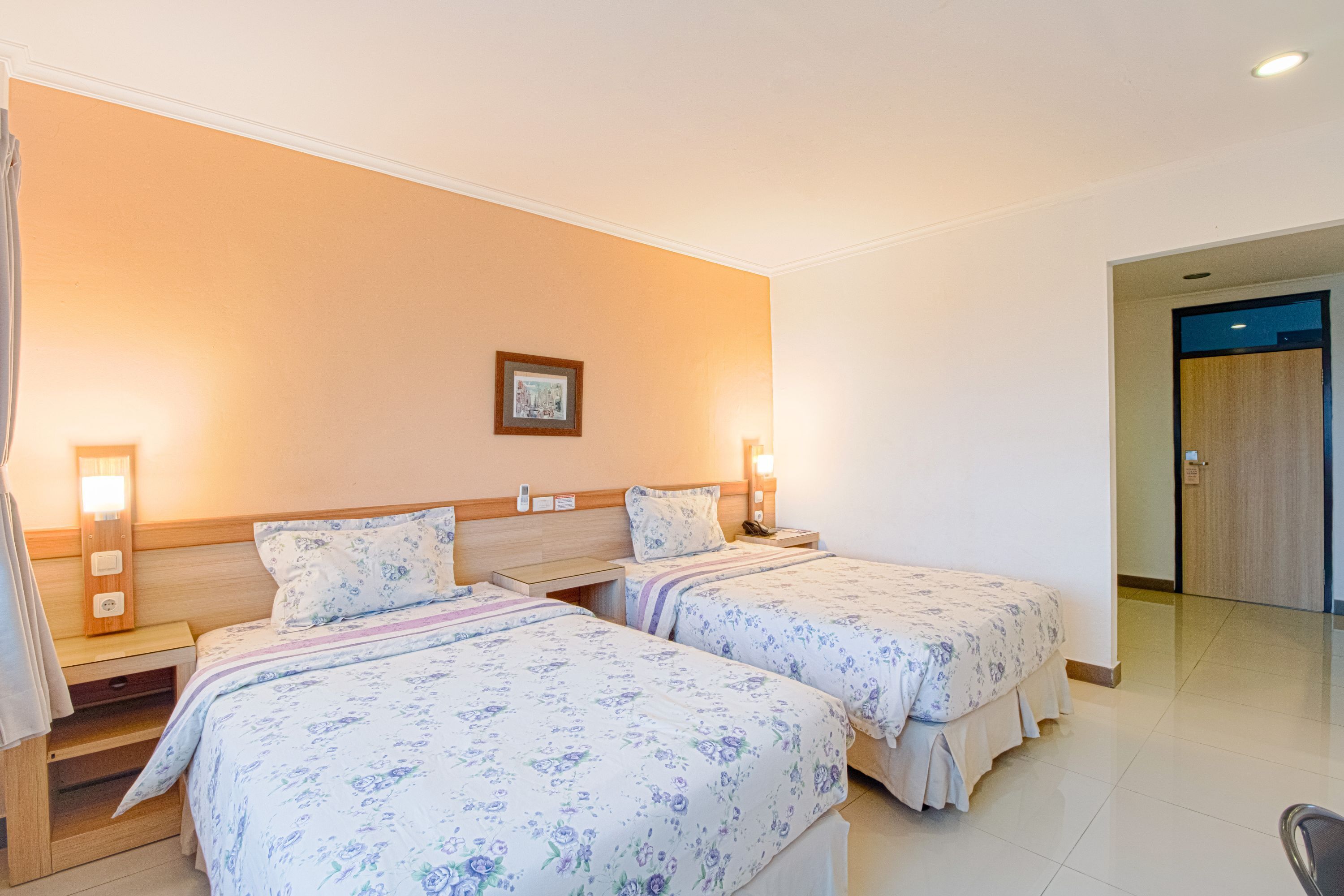 Une chambre d’hôtel avec deux lits, chacun recouvert d’une couvre-lit à motifs floraux, dotée de lampes de chevet et ornée d’un tableau encadré au mur.