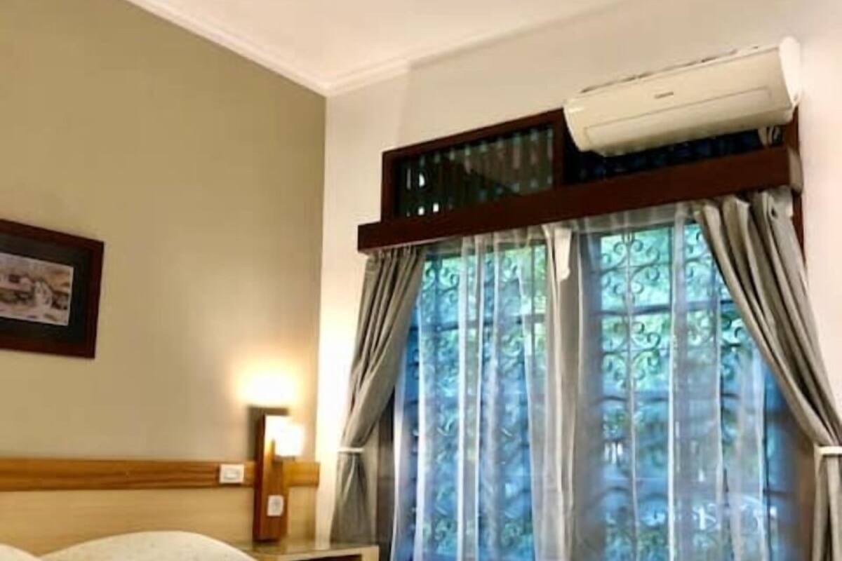 A hotel room with a large bed, a nightstand, a wall-mounted air conditioner, and a window with curtains.