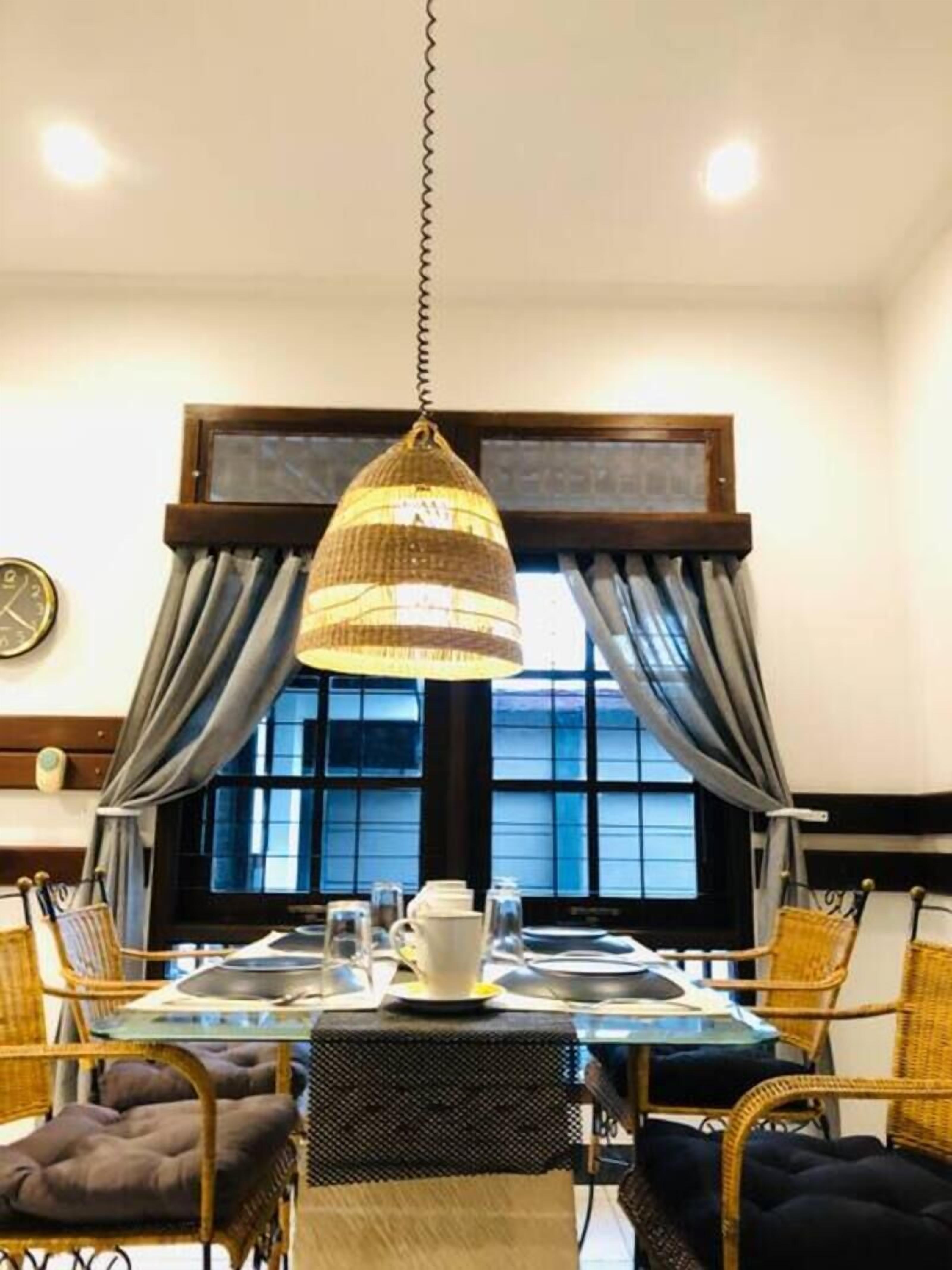 A dining area with a table set for four, surrounded by wicker chairs. There is a hanging lamp above the table, and a clock on the wall to the left.