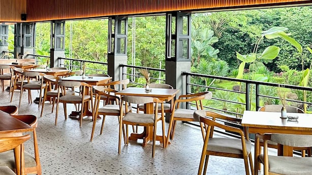 A spacious indoor area with wooden ceiling panels and ceiling fans, large windows offering a view of greenery outside, and wooden tables and chairs arranged for dining.