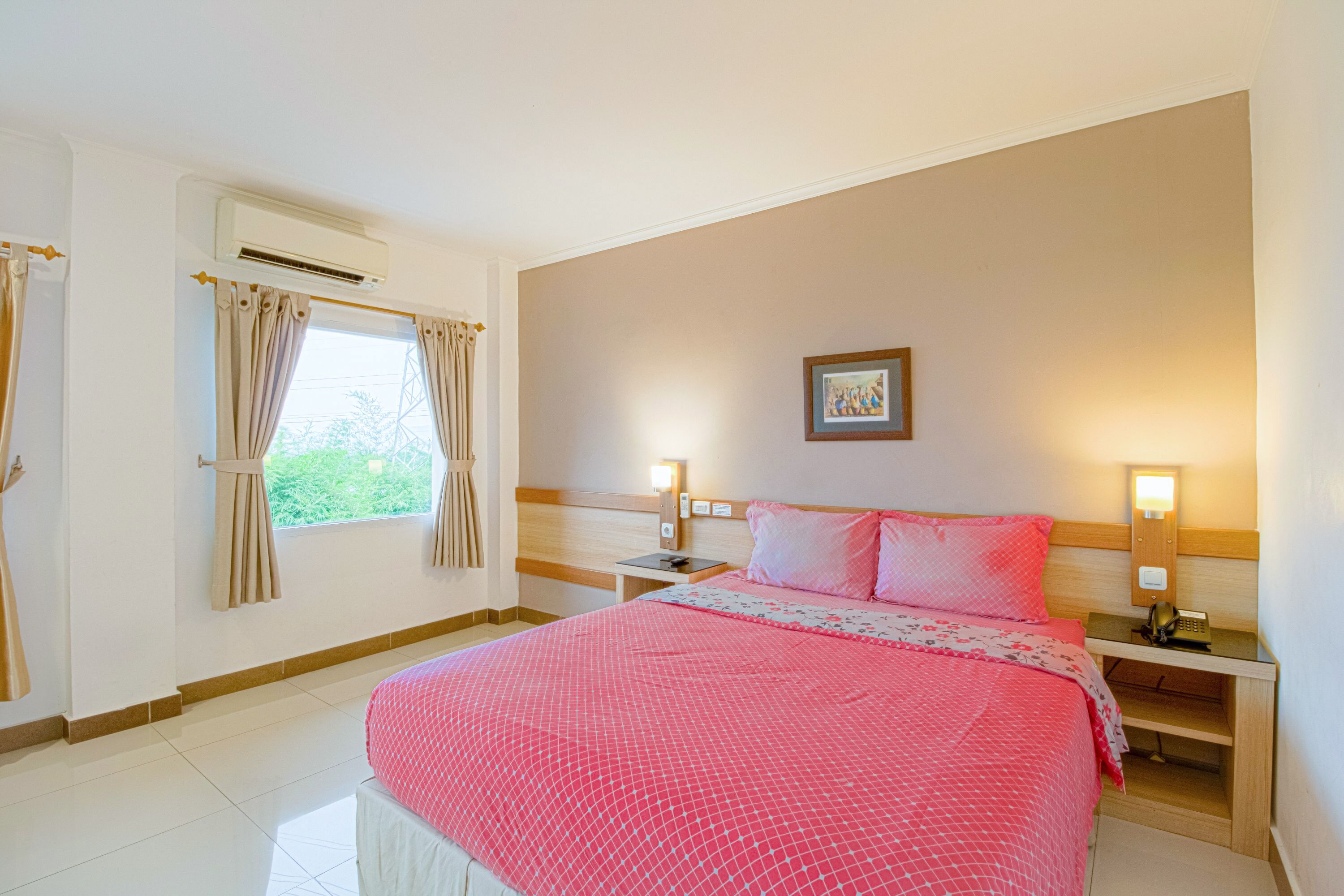 A hotel room with a large bed, two bedside tables, a nightstand, an air conditioning unit, a window with curtains, and a wall-mounted picture.