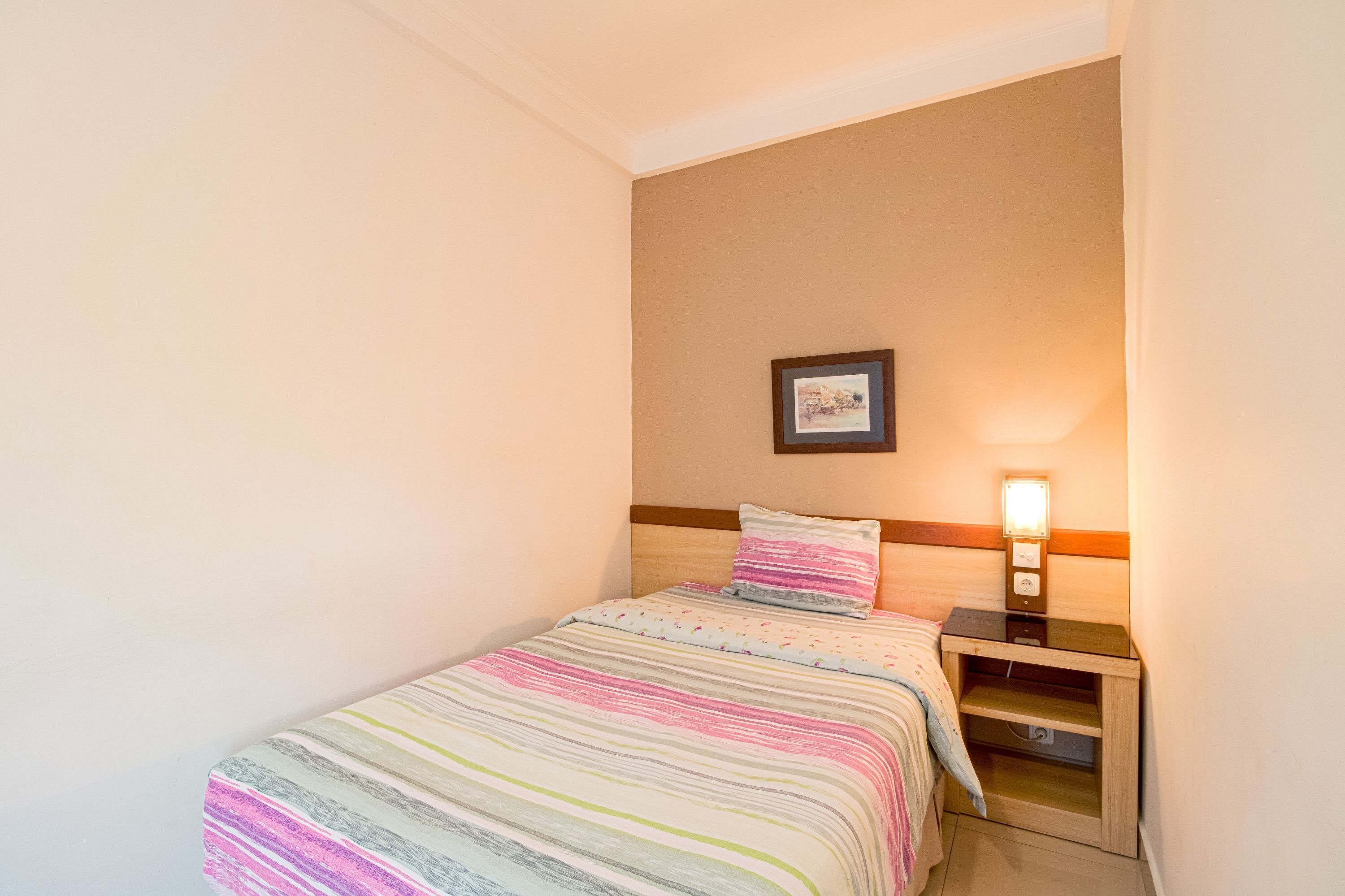 A small, neatly arranged bedroom with a single bed, a bedside table with a lamp, and a framed picture on the wall.