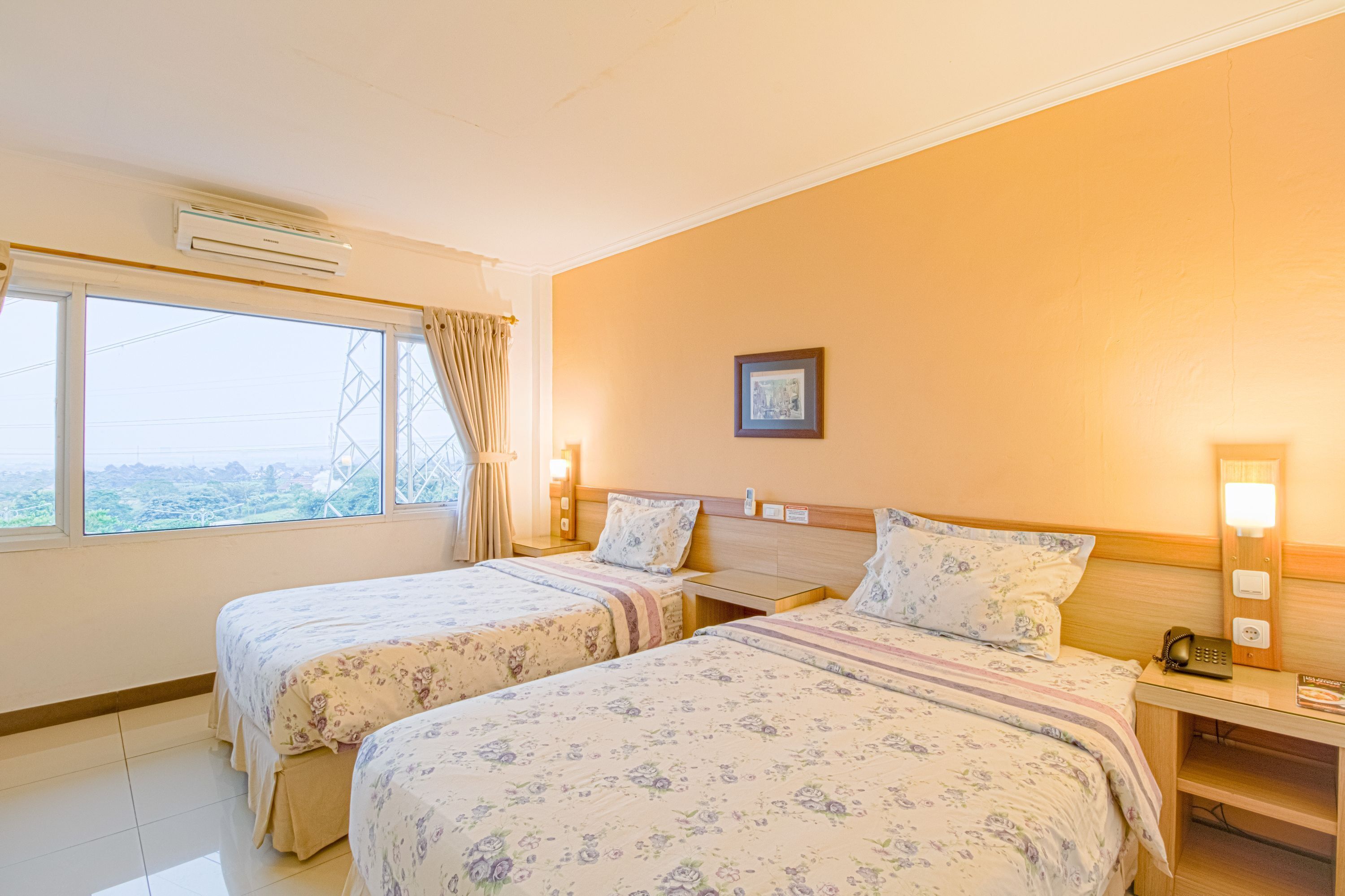 Ein Hotelzimmer mit zwei Betten, einem großen Fenster mit Vorhängen, einer Klimaanlage und einem Nachttisch mit Lampe und Telefon.