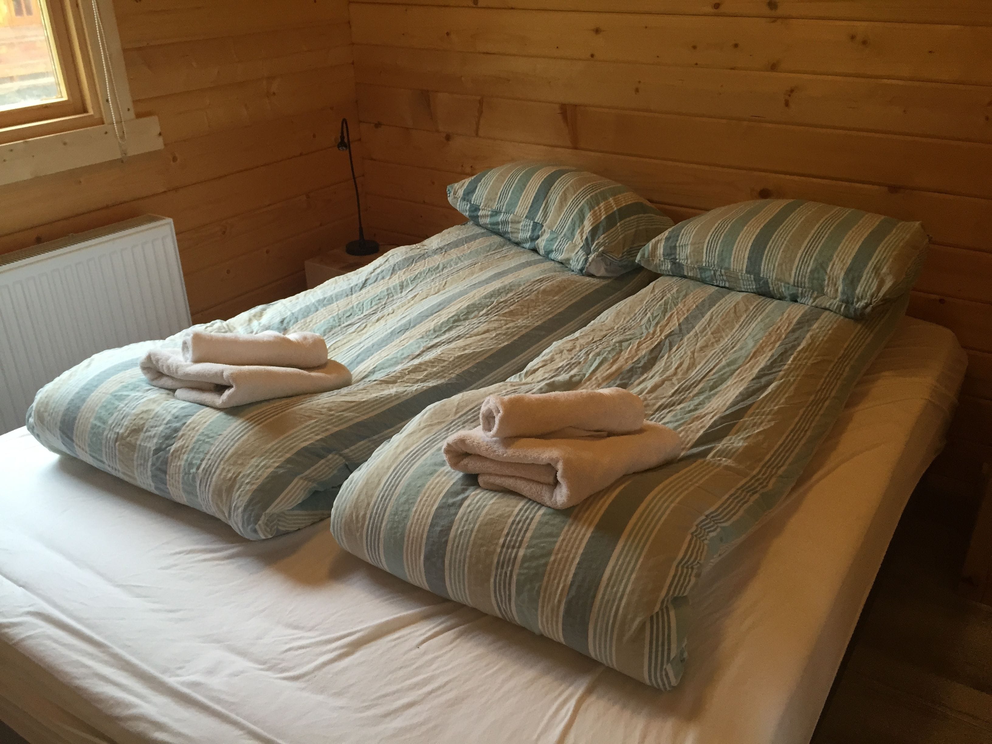 A double bed with striped bedding and two pillows, a radiator, and a window with a chain.