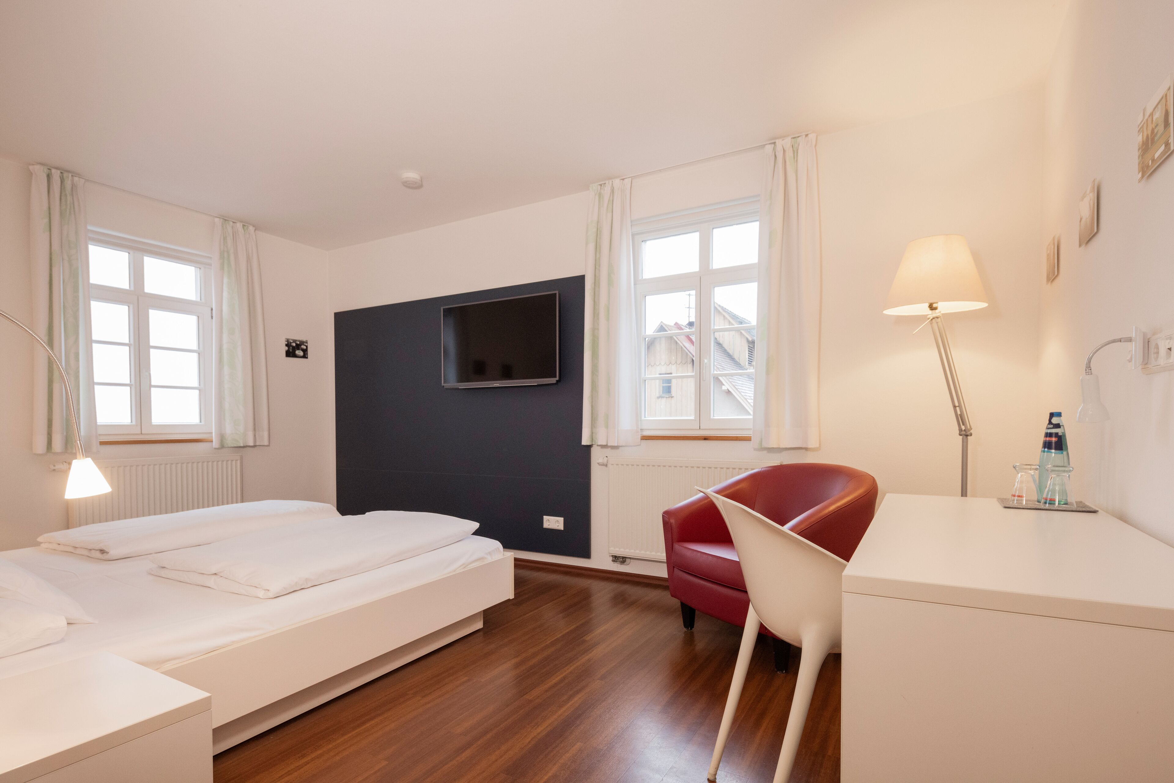 A hotel room with a bed, a desk, a chair, a TV mounted on the wall, and a window with curtains.
