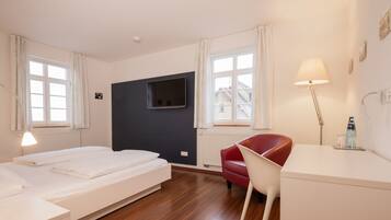 A hotel room with a bed, a desk, a chair, a TV mounted on the wall, and a window with curtains.