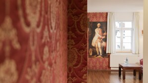 A room with a large painting on the wall, a window with curtains, a wooden table, and a red patterned carpet. - Brauerei Gasthof Krone (Tettnang)