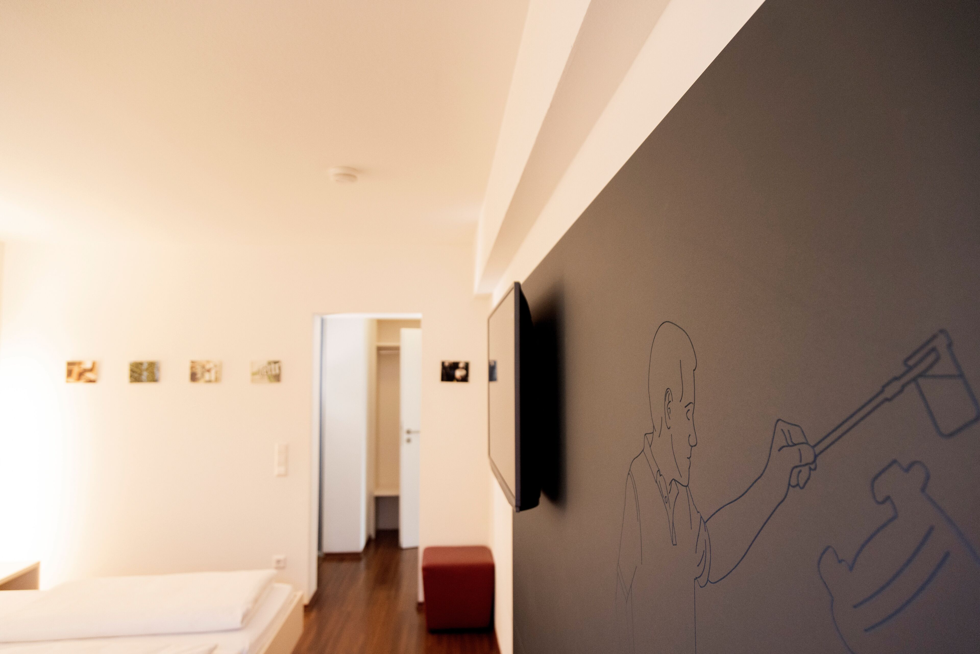 A hotel room with a bed, a red ottoman, a wall-mounted TV, and a hallway leading to another room.