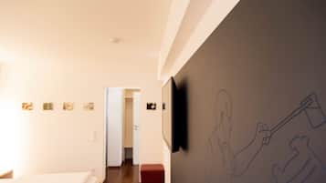 Une chambre d’hôtel avec un lit, un pouf rouge, un téléviseur fixé au mur et un couloir menant à une autre pièce.