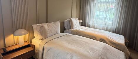 A hotel room with two beds, a nightstand with a lamp, a window with curtains, and a ceiling light.