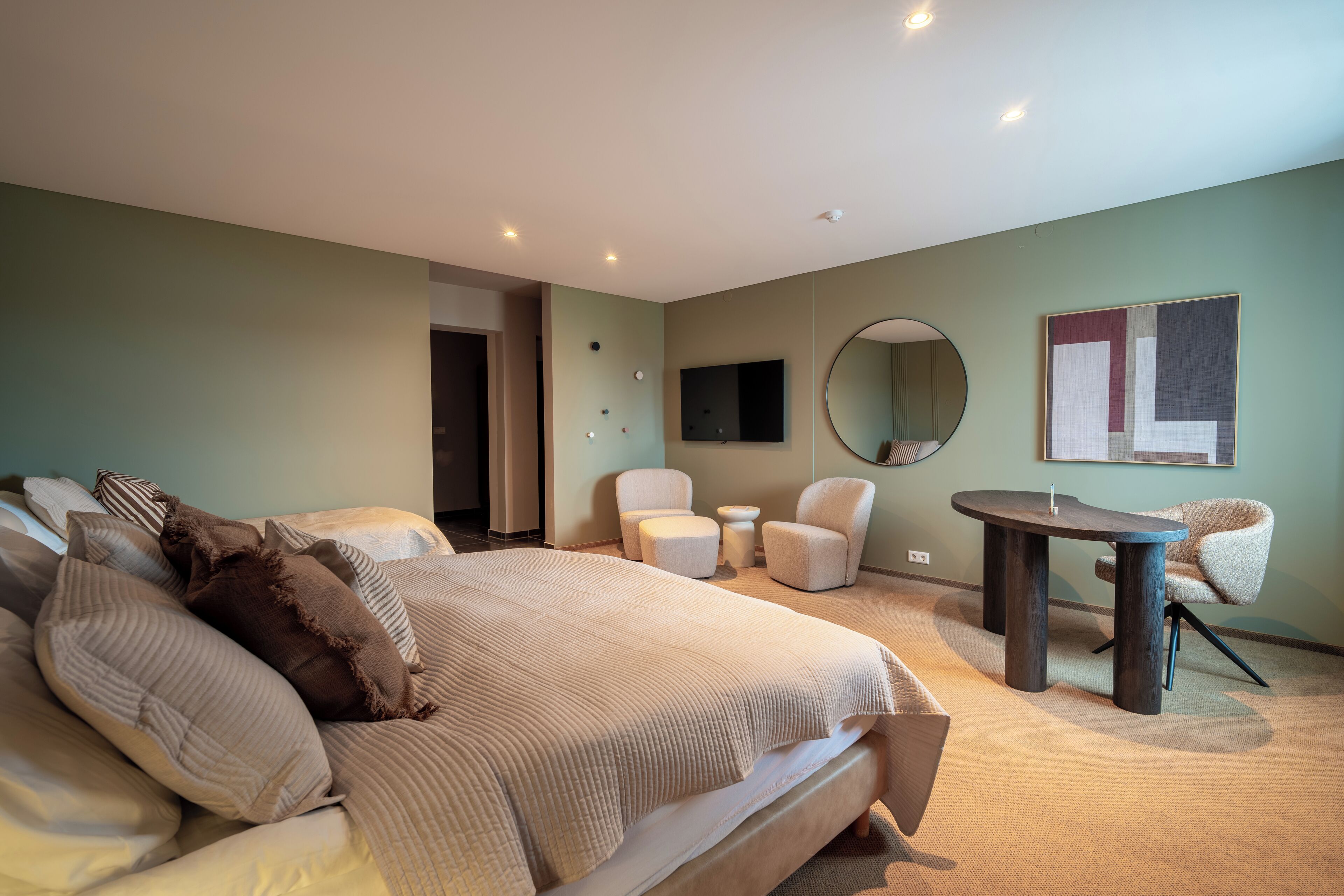 A modern hotel room with a large bed, two armchairs, a small table, a round mirror, and a flat-screen TV mounted on the wall.