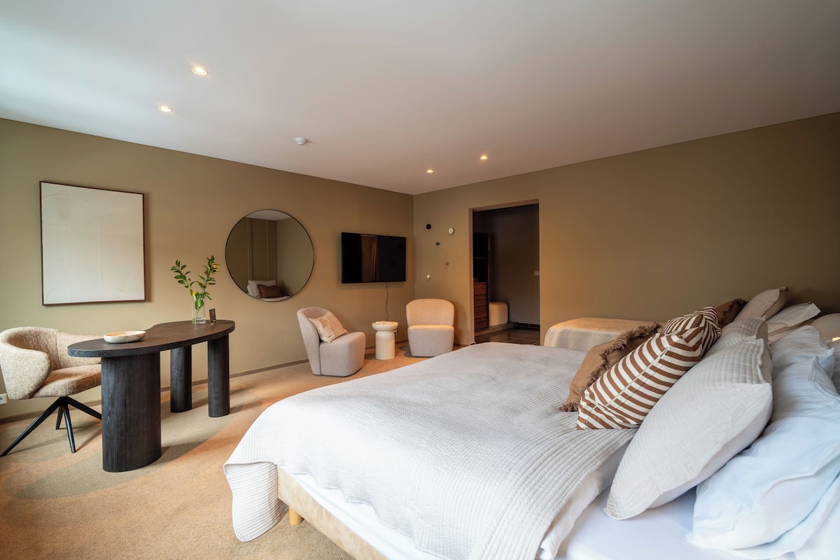 Un dormitorio moderno con una cama grande, un espejo redondo, dos sillones, una mesita y un televisor en la pared.