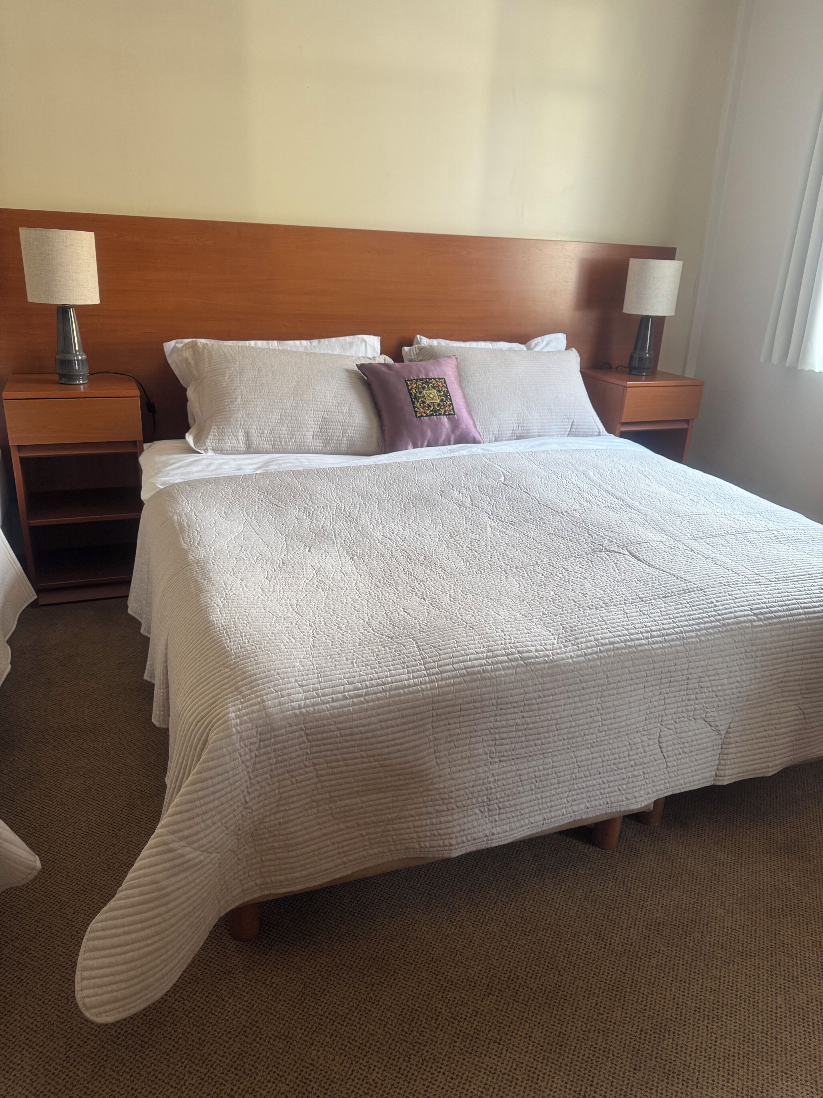 Una cama bien tendida con un edredón blanco, dos mesitas de noche con lámparas y un cabecero de madera.