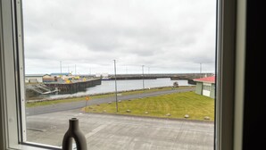 Une fenêtre ornée d’un vase posé sur le rebord, donnant sur un port avec un pont, une zone herbeuse et un bâtiment avec un toit rouge.