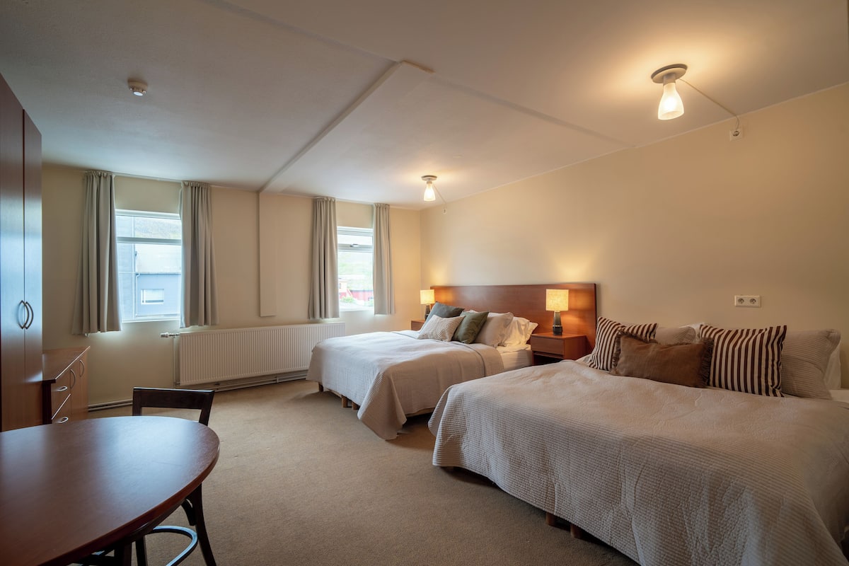 Habitación de hotel con dos camas, una mesa de madera y un ventanal con cortinas.
