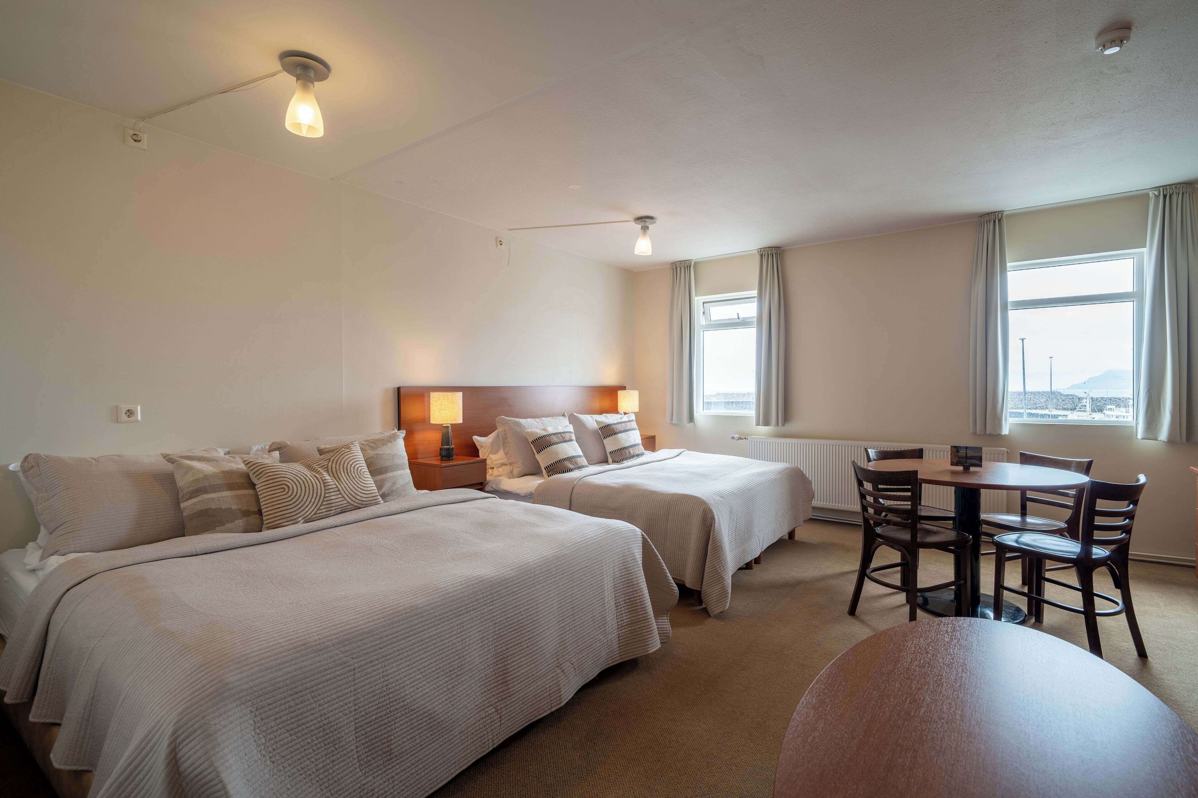 Une chambre d’hôtel avec deux lits, une table à manger et des chaises. Il y a une fenêtre avec des rideaux, un luminaire au plafond et un radiateur.
