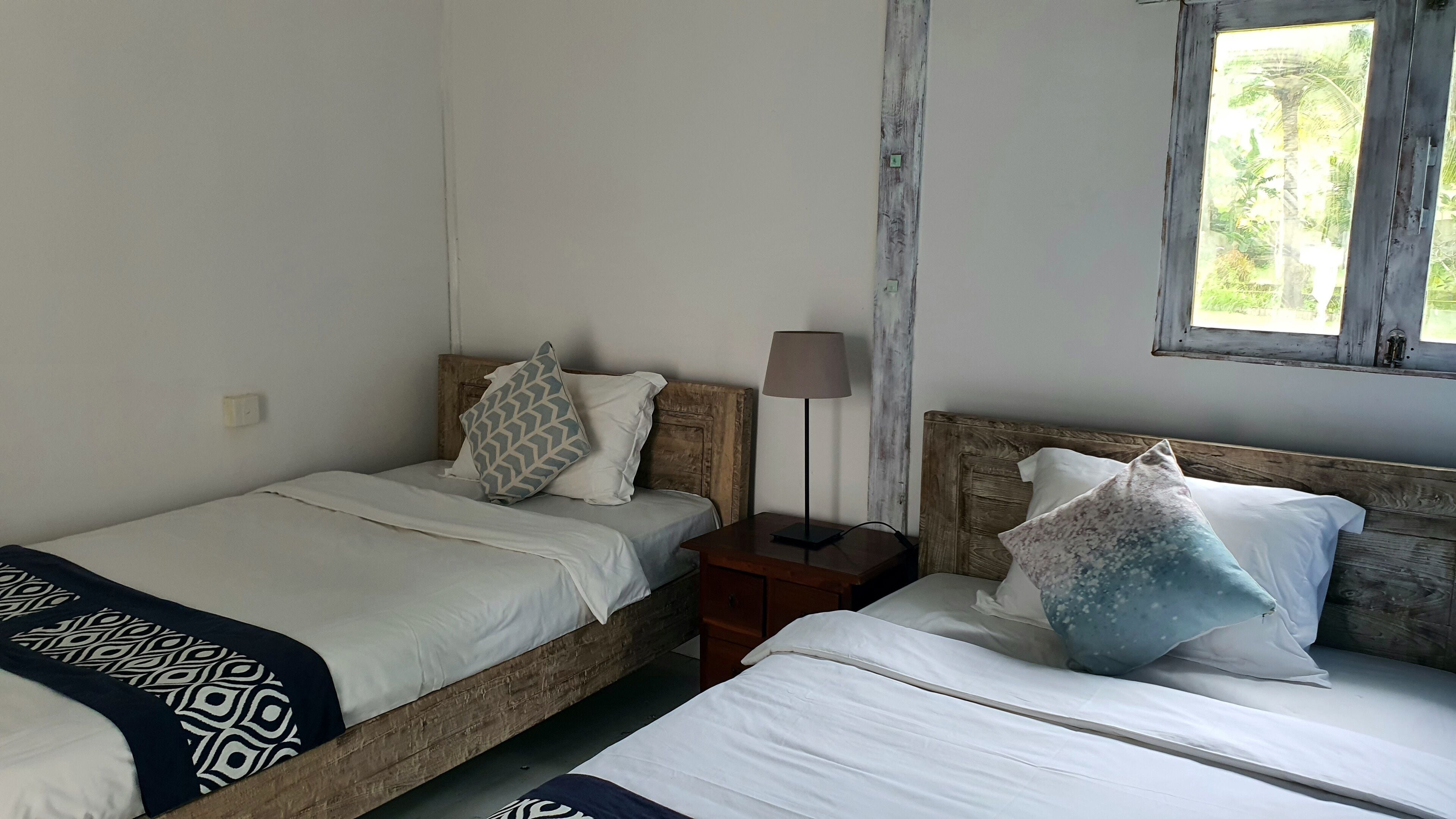 A room with two single beds, a nightstand with a lamp, and a window with a view of greenery.
