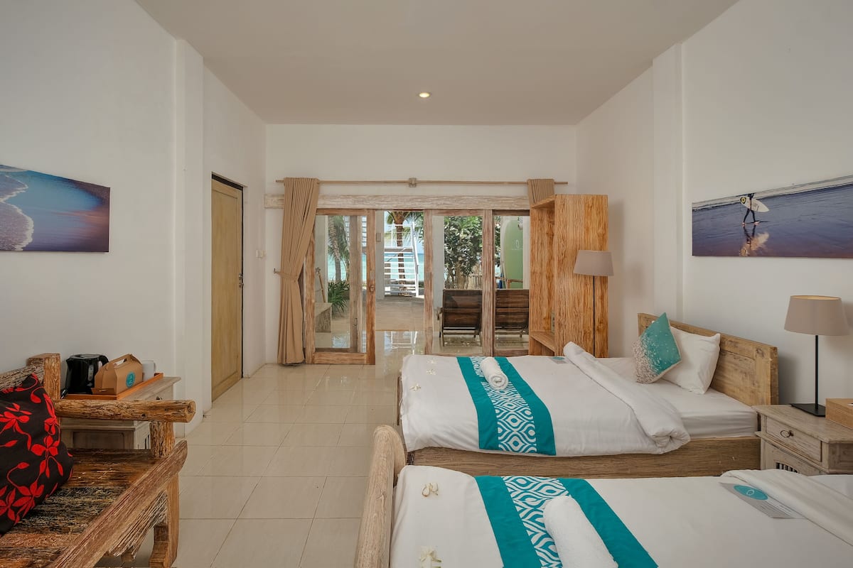 A hotel room with two beds, a wooden nightstand, a desk with a lamp, and a view of the outdoor area through glass doors.