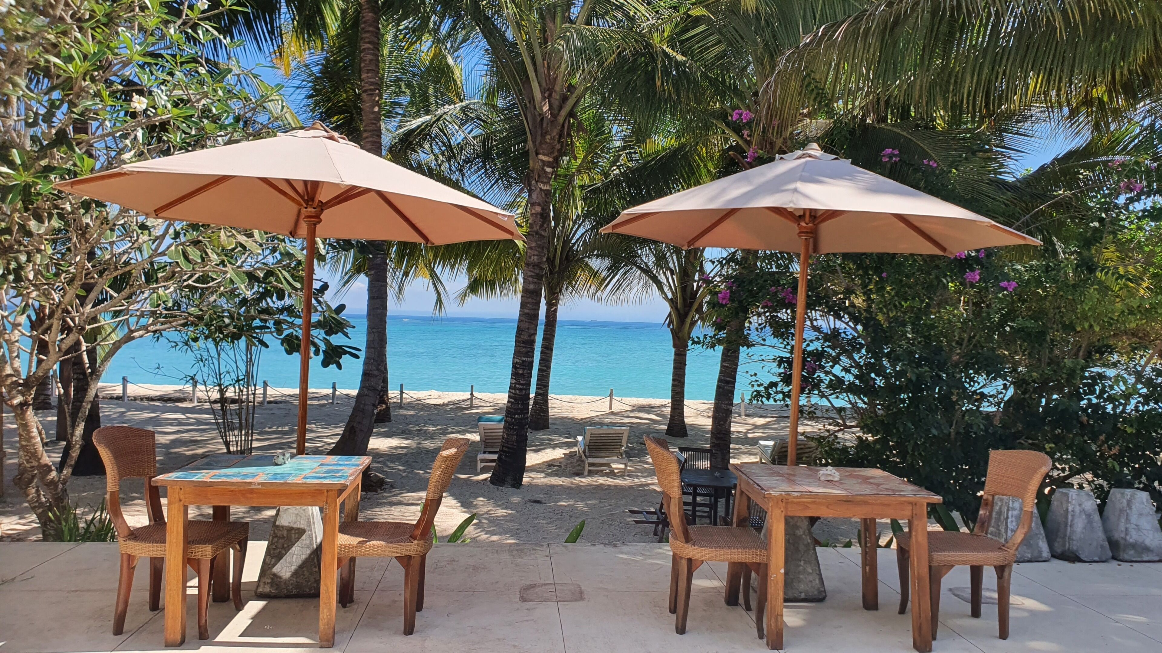 A beachside patio with two wooden tables and chairs, each under a beige umbrella, surrounded by palm trees and a view of the ocean.