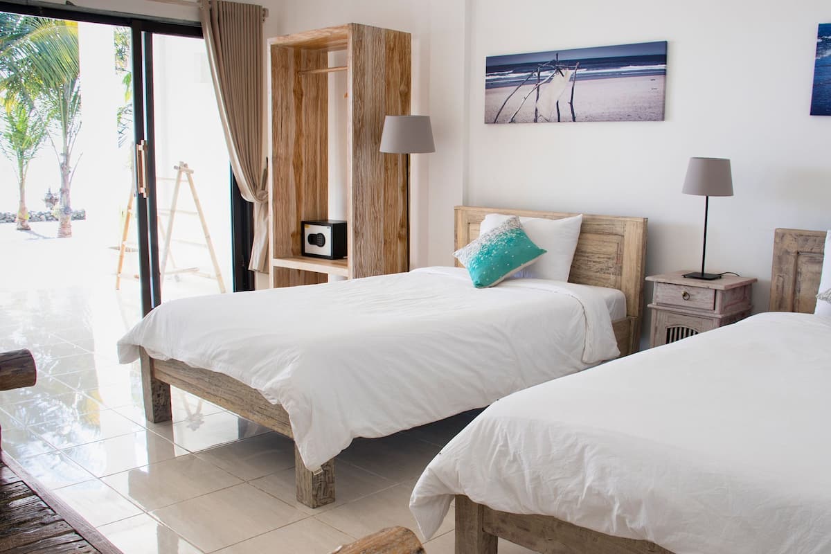 A room with two beds, a wooden cabinet, a beach painting, and a lamp.