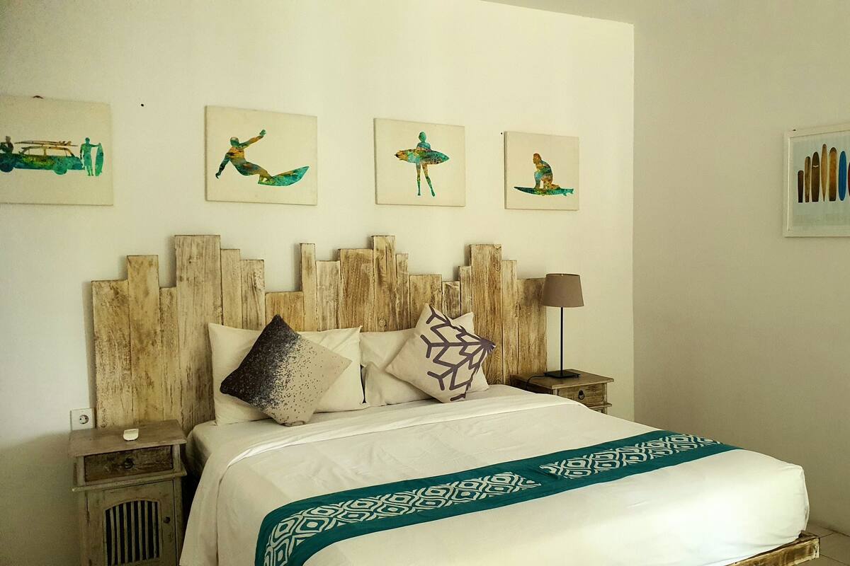 A neatly arranged bedroom with a wooden headboard, a bed with patterned bedding, a bedside table with a lamp, and three wall-mounted artwork depicting surfers.