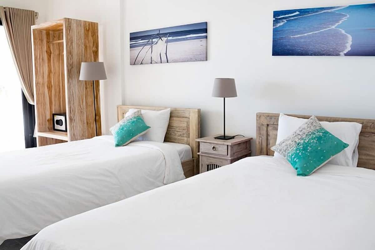 Two beds with white bedding and teal pillows, a wooden nightstand with a lamp, and a beach-themed picture on the wall.