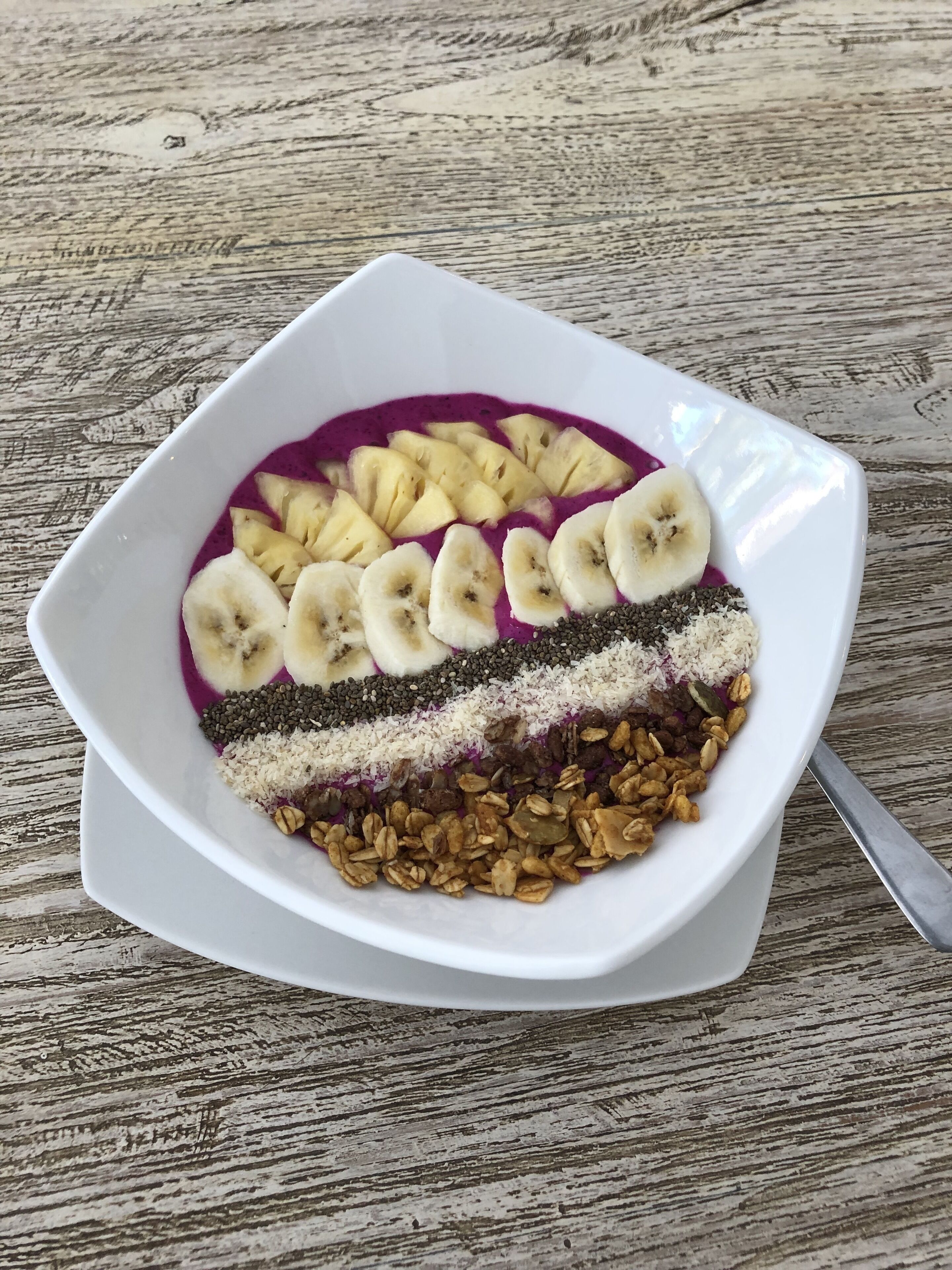 Un tazón blanco con un desayuno en capas que incluye rodajas de plátano, semillas de chía, granola y una base de batido morado.