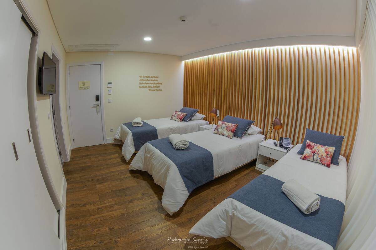 A hotel room with three beds, wooden flooring, a TV, and a wall-mounted light fixture.