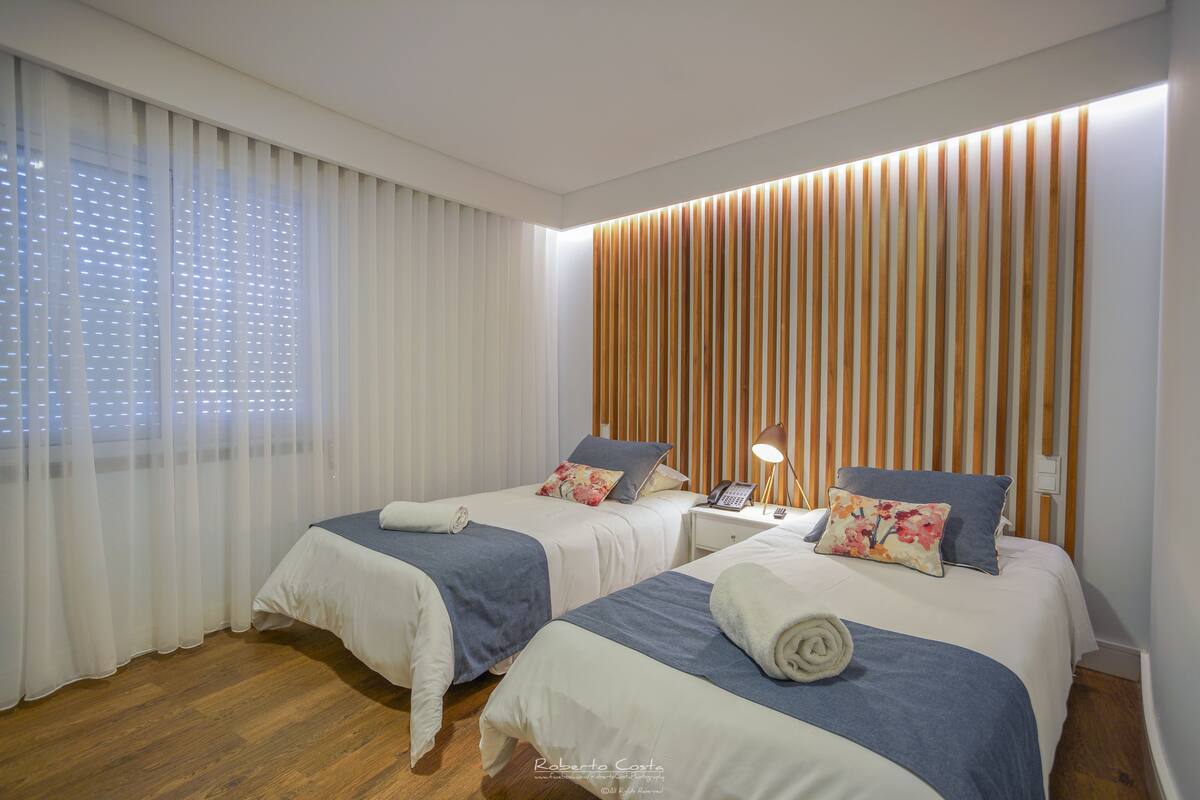 A hotel room with two beds, a wooden slat wall, a nightstand with a lamp, and a window with sheer curtains.