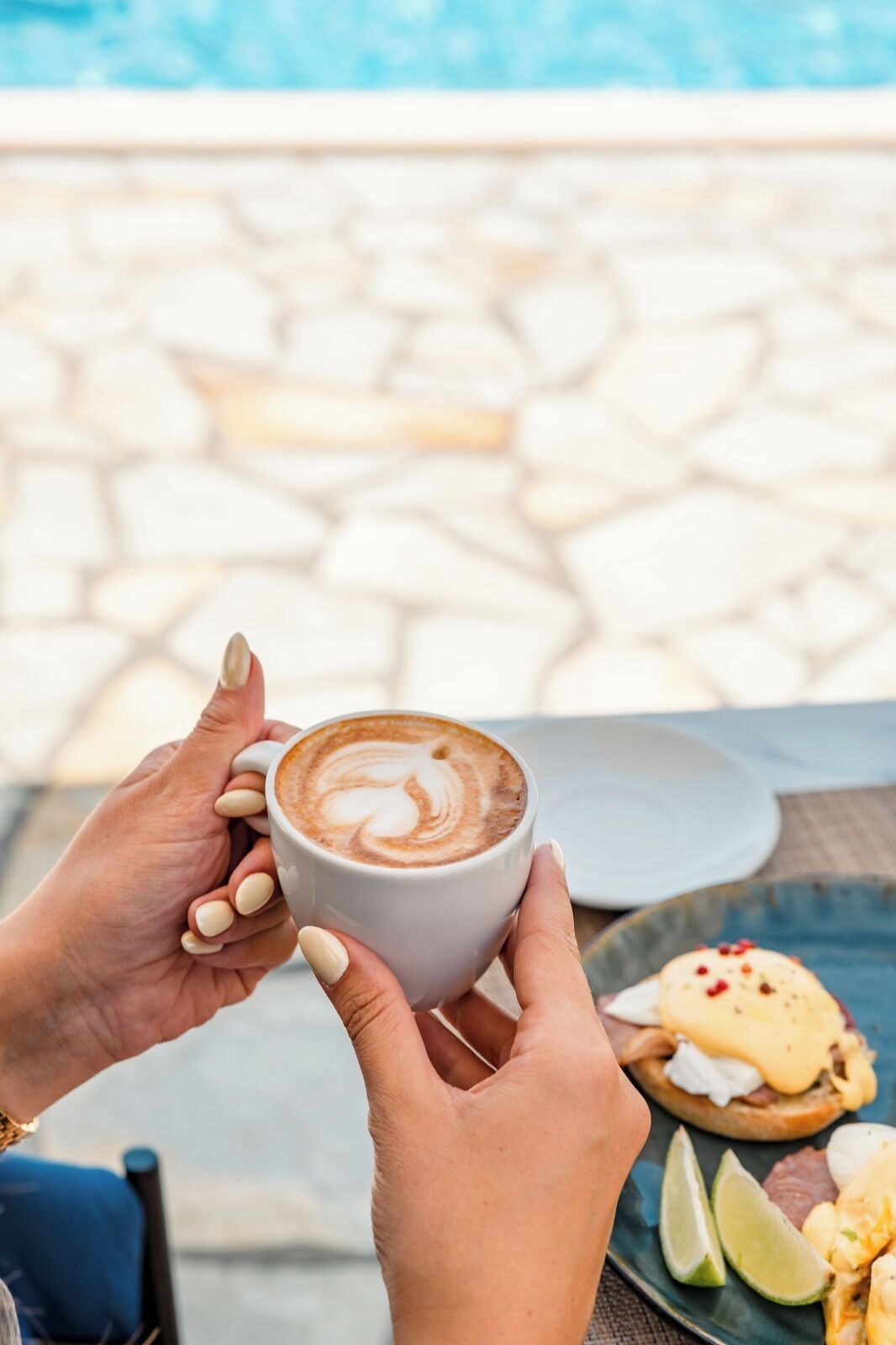 A person holding a cup of coffee with latte art, a plate with food, and a pool in the background.
