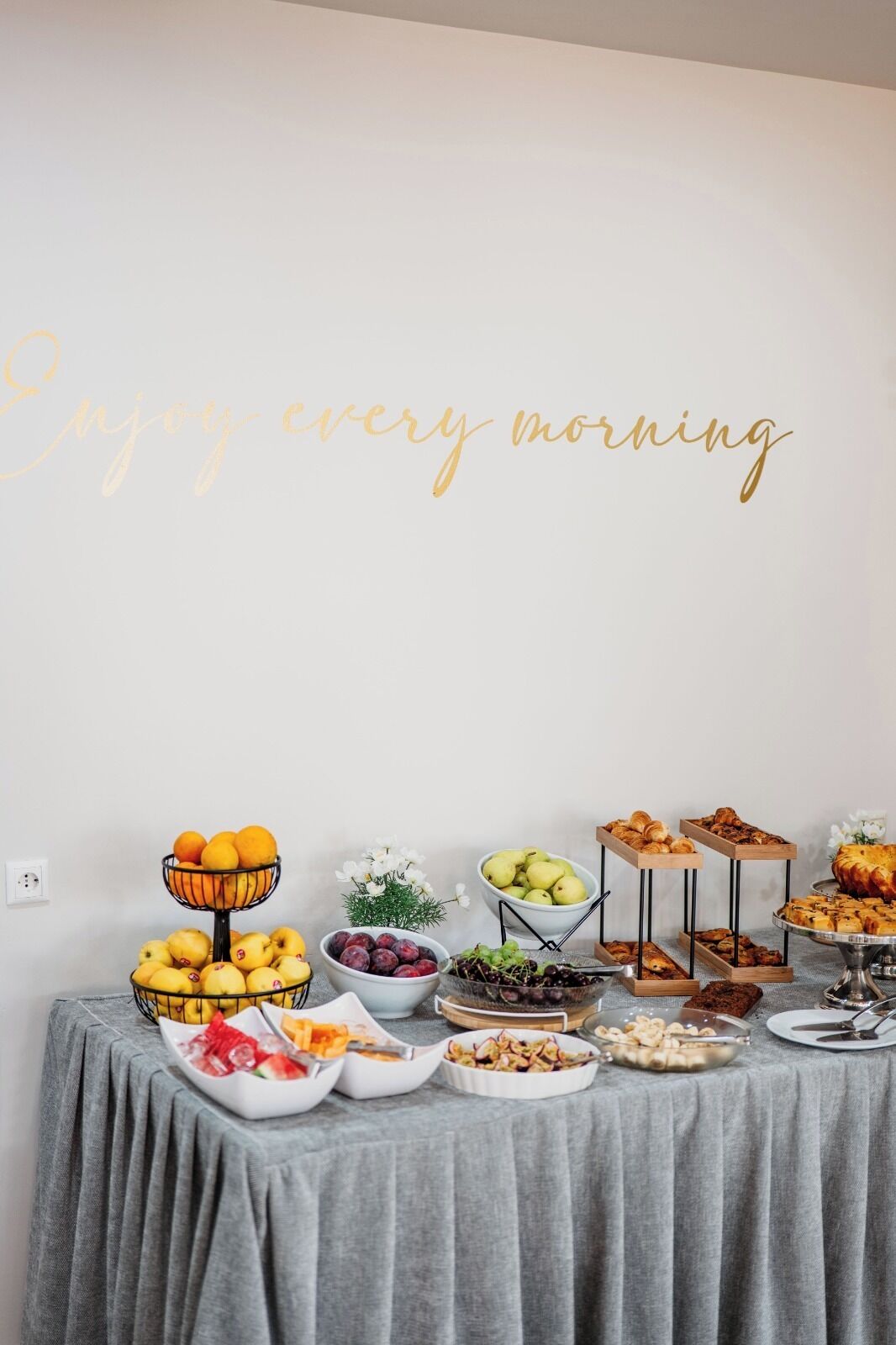 A breakfast buffet with various fruits, pastries, and a decorative sign saying 'Enjoy every morning.'