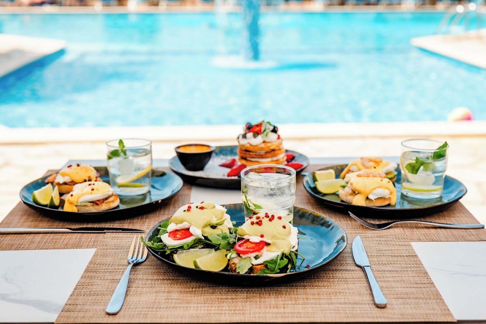 A dining table with plates of food and drinks, a pool in the background.
