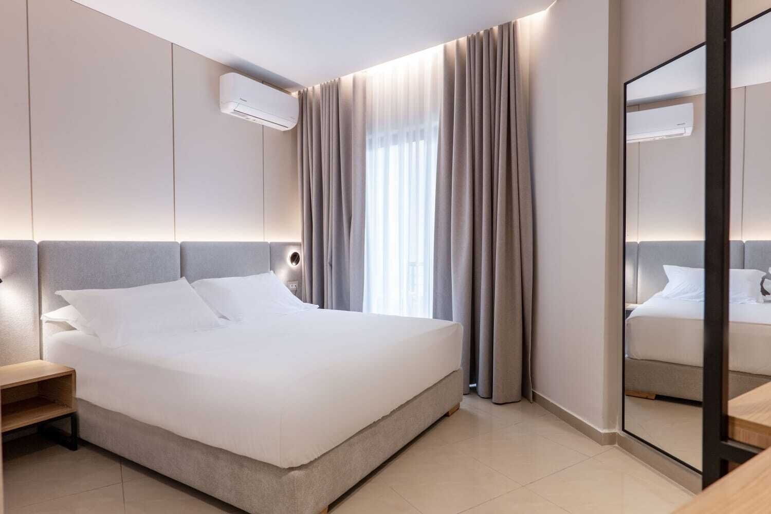 A modern hotel room with a large bed, a bedside table, an air conditioning unit, and a mirror.