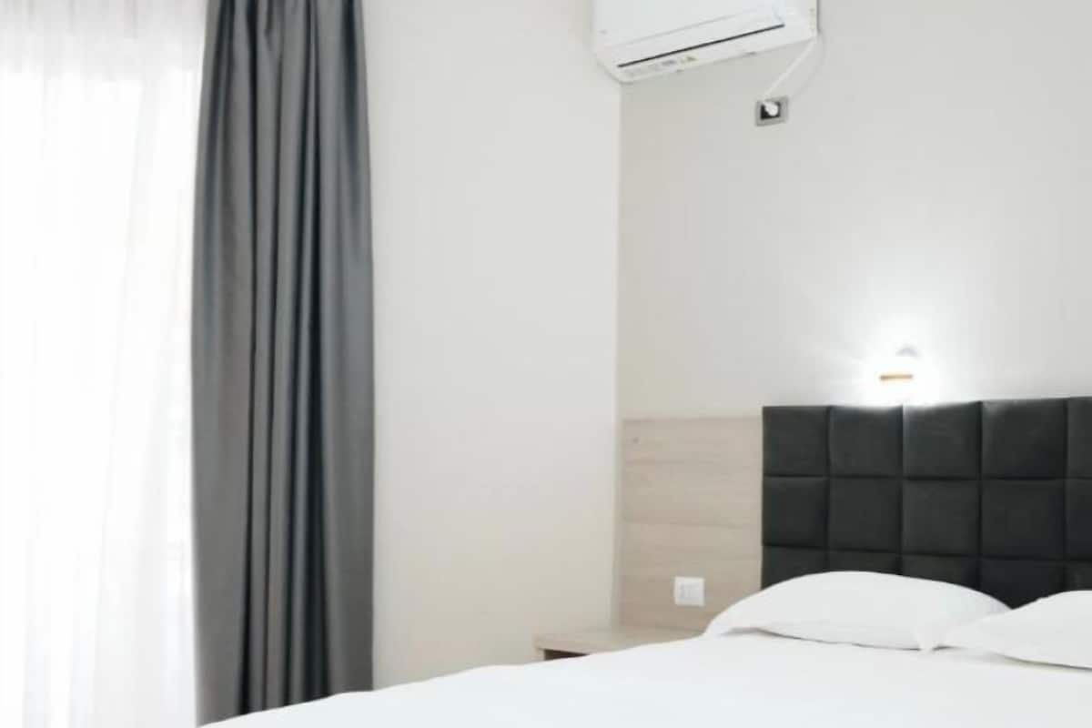 Habitación de hotel con cama, cabecero, unidad de aire acondicionado y ventana con cortinas.