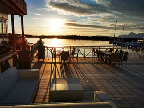 A deck with outdoor seating, overlooking a calm body of water and boats, with mountains in the background. - Kjerringøy Bryggehotell (Bodo)