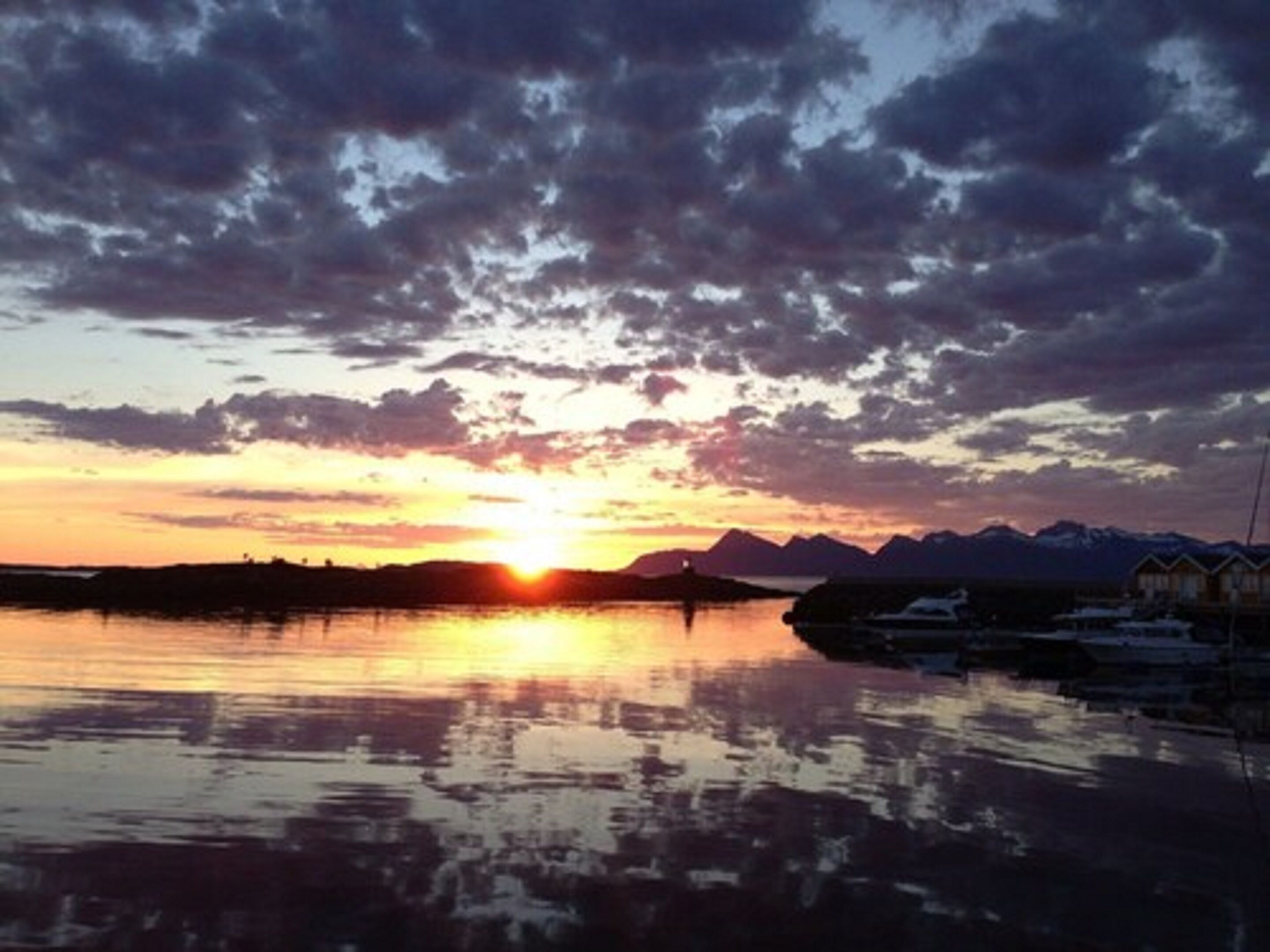 A sunset over a calm body of water with mountains in the background and a row of cabins along the shore.