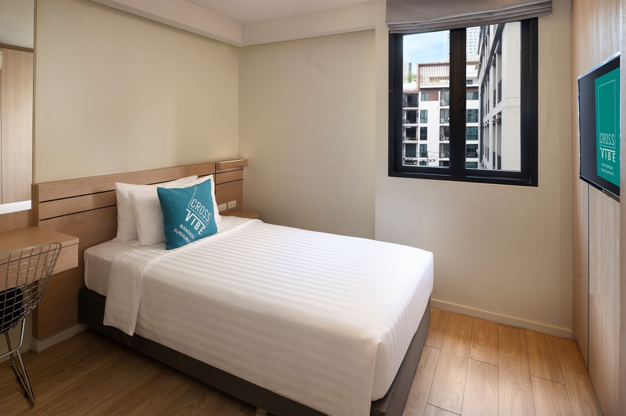 Een netjes opgemaakt bed met een witte matras en een blauwgroen kussen, een houten nachtkastje, een metalen stoel en een raam met uitzicht op gebouwen.