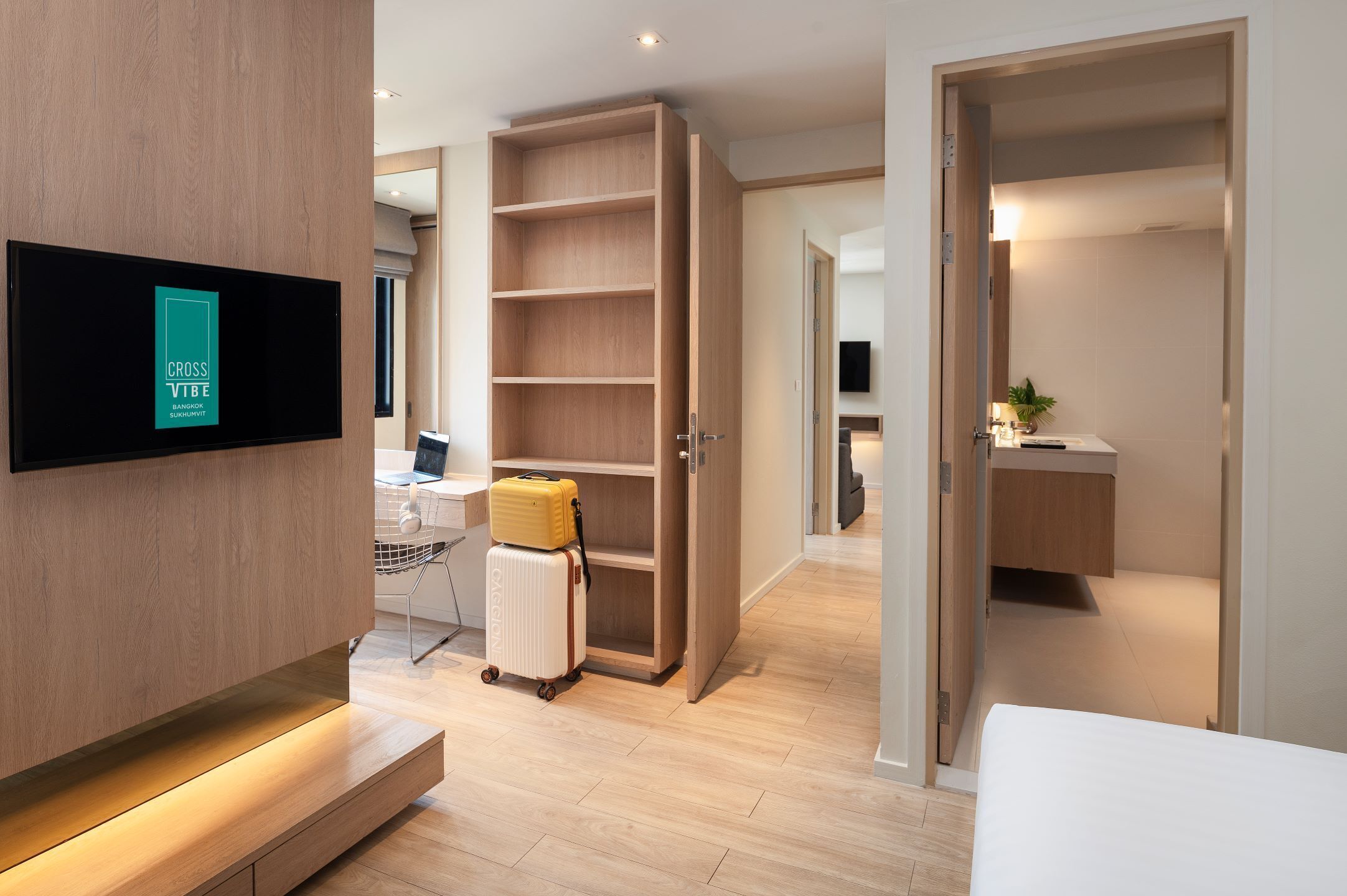 A modern hotel room with a wooden wardrobe, a flat-screen TV mounted on the wall, a desk with a computer, and a suitcase on the floor.