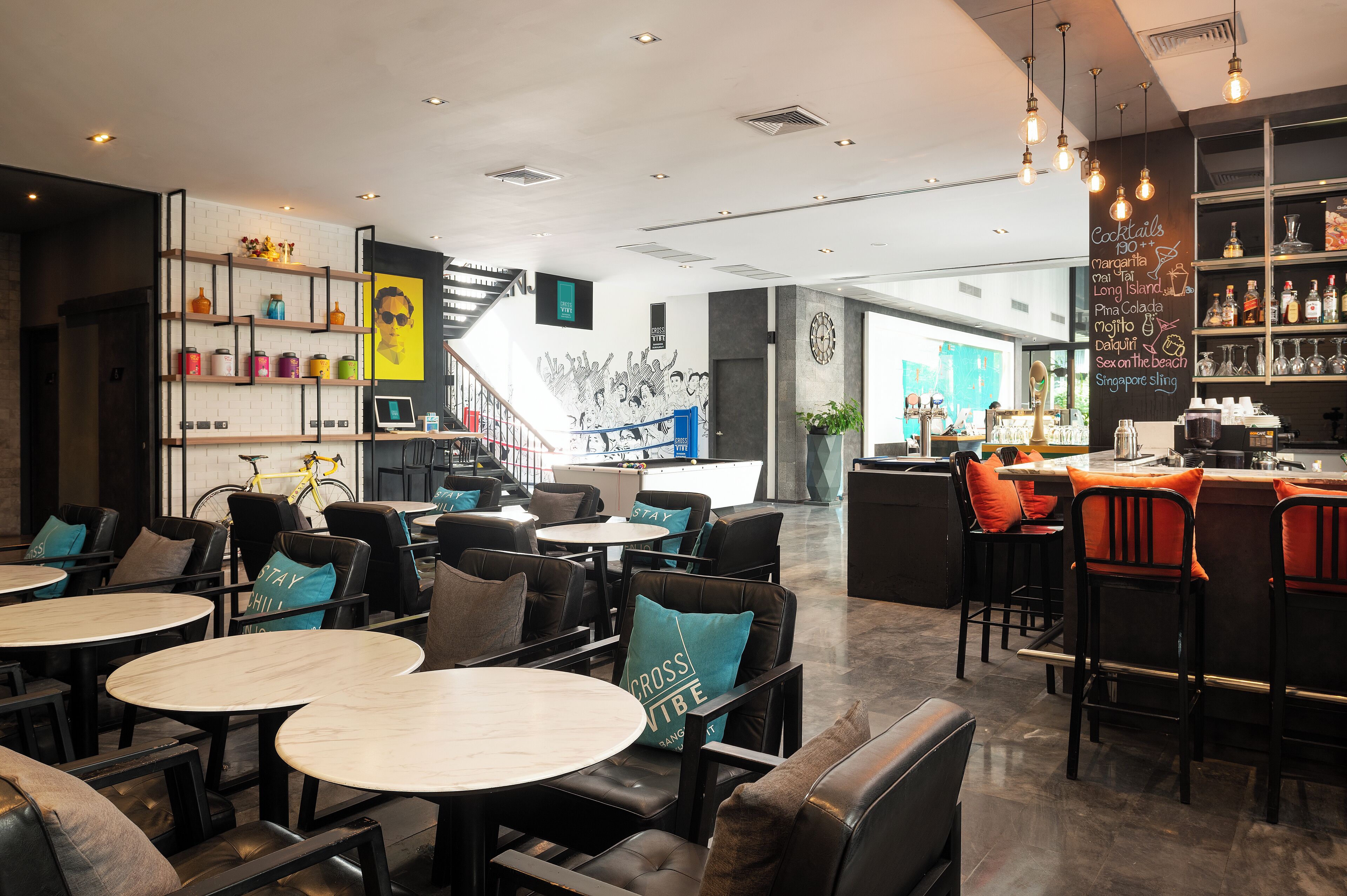 Un espace bar moderne avec des sièges, un escalier et un mur où est affichée la carte des cocktails.
