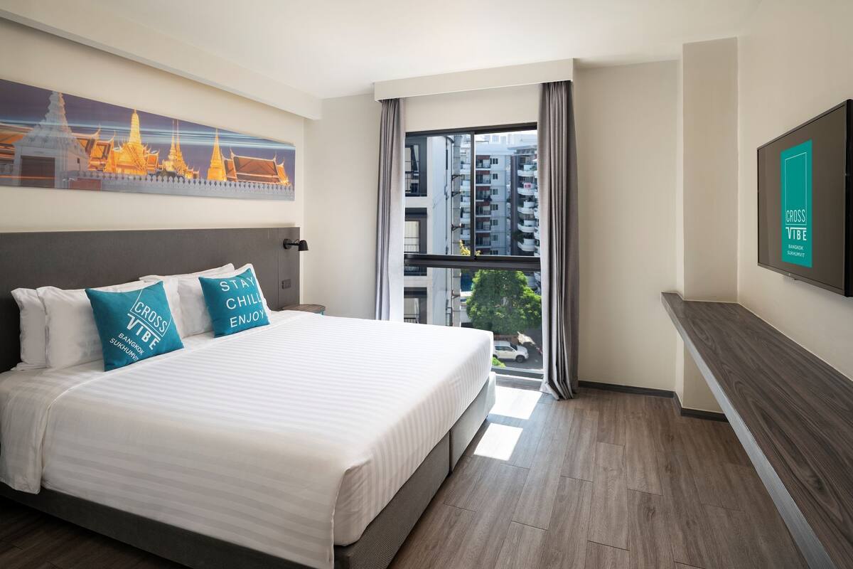Ein modernes Hotelzimmer mit großem Bett, Blick auf die Stadt durch ein Fenster und einem an der Wand befestigten Fernseher.