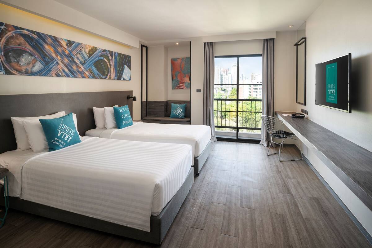 Ein Hotelzimmer mit zwei Betten, einem Fernseher, einem Schreibtisch und einem großen Fenster mit Blick auf die Stadt.