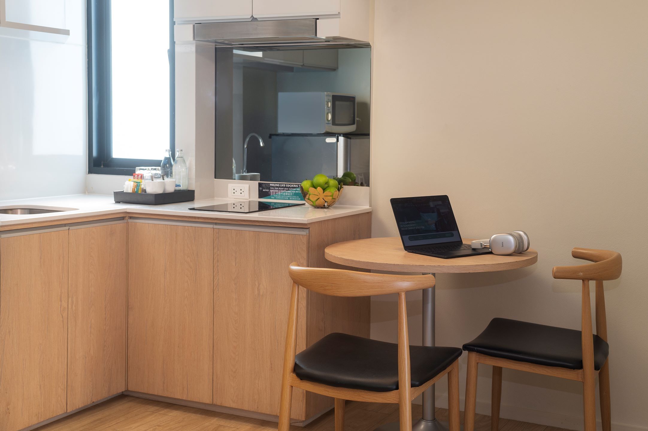 A compact kitchen with wooden cabinets, a microwave, and a laptop on a small round table. There are two wooden chairs with black cushions.