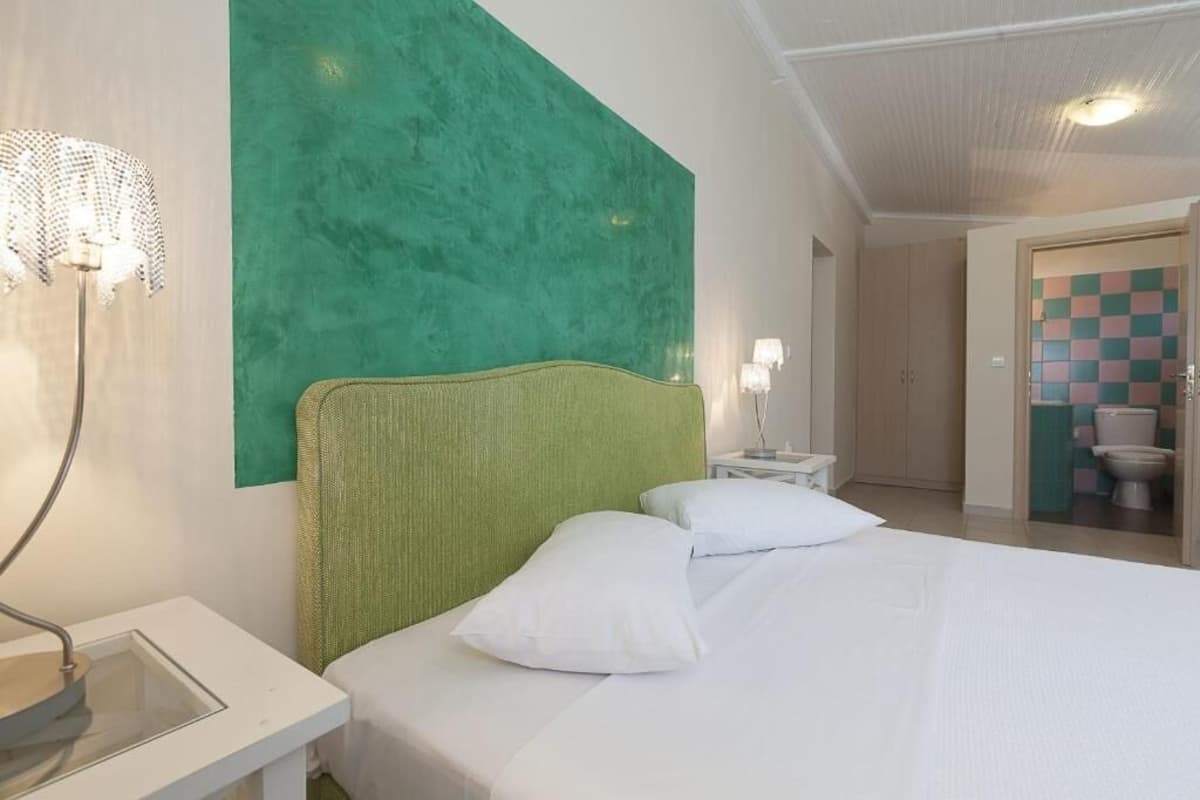 Quarto de hotel com uma parede verde de destaque, cama com roupa de cama branca, mesa de cabeceira com abajur e vista para o banheiro.