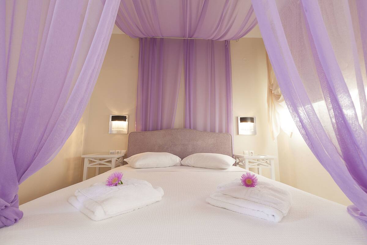 A bedroom with a large bed, white bedding, and purple drapes. There are two bedside tables with lamps.