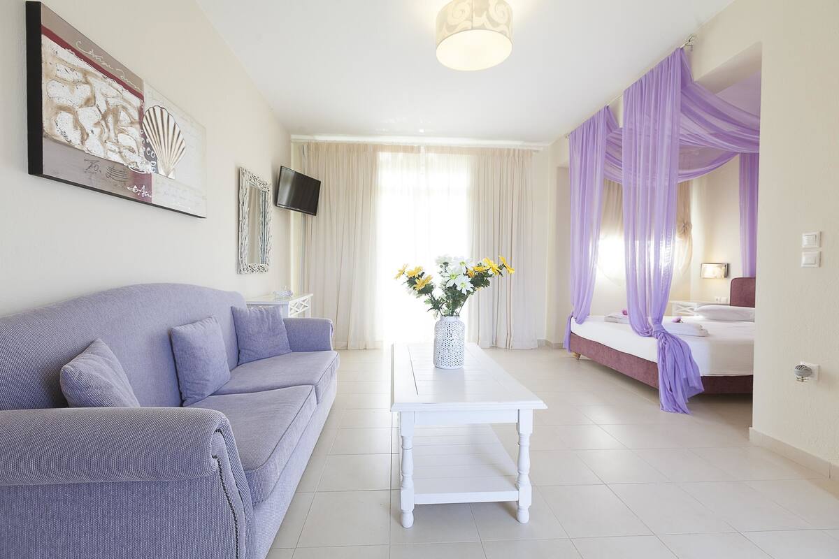 A modern hotel room with a sofa, a white coffee table, a bed with purple curtains, and a TV mounted on the wall.