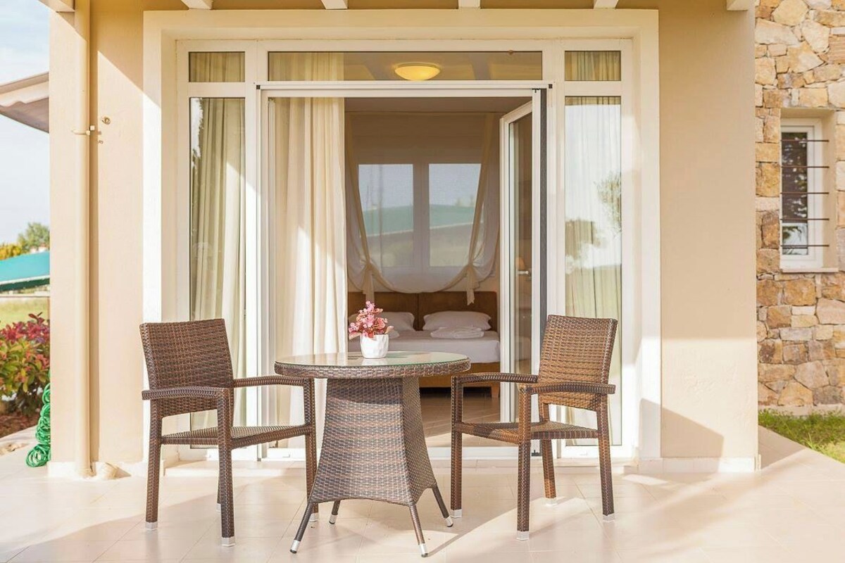 An open sliding glass door leading to a bedroom with a bed, a small table with a flower vase, and wicker chairs.