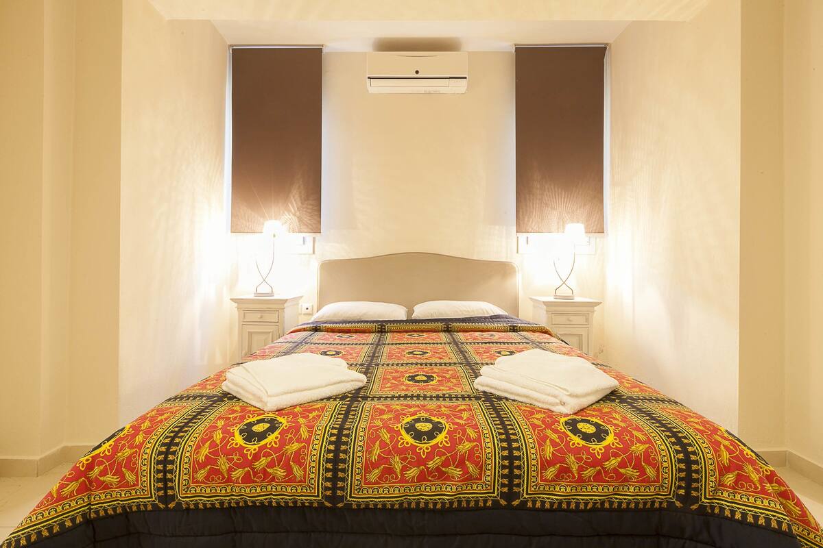 A neatly made bed with a patterned bedspread, two bedside tables with lamps, and an air conditioning unit on the wall.